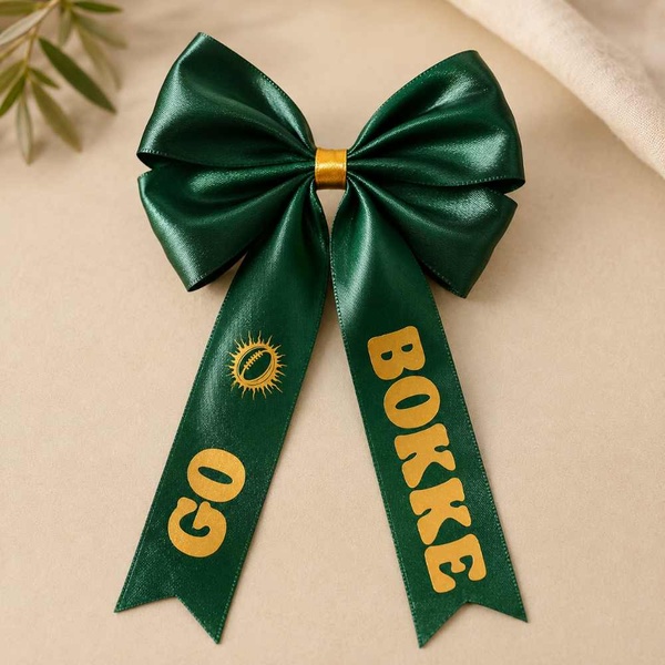 Emerald green satin Go Bokke hair bow with gold print and ribbon tails, displayed on a warm neutral background in a polished product photo.