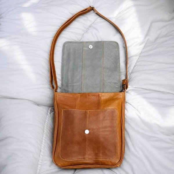 Tan leather crossbody messenger bag with flap closure and adjustable strap, shown open on white bedding with gray interior lining.