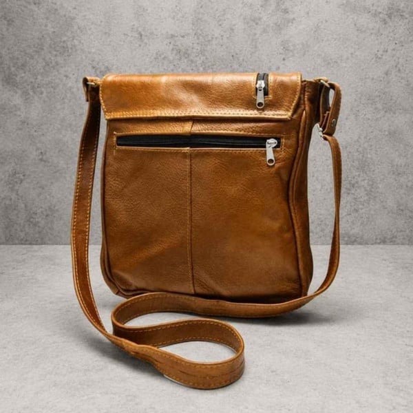 Tan leather crossbody bag with multiple zip pockets and adjustable strap displayed on a neutral concrete background.
