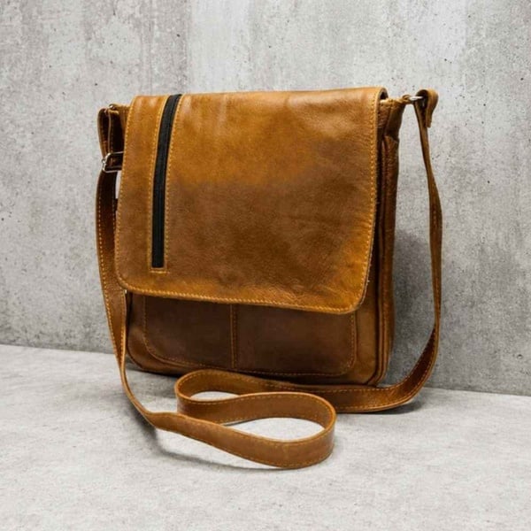 Tan leather crossbody messenger bag with flap closure and vertical zip pocket, styled on a textured concrete backdrop.