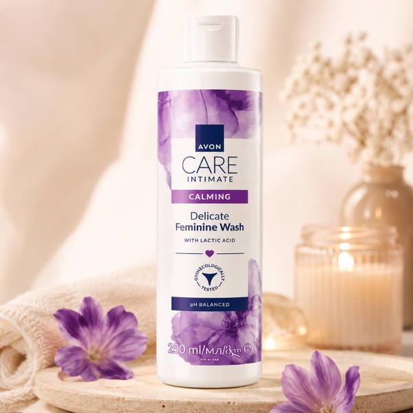Avon Care Intimate Calming Delicate Feminine Wash 250ml with lactic acid on a warm spa-inspired setting with purple flowers.